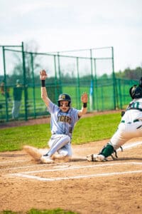 Baseball player sliding into home plate with hands raised, wearing a Tigers jersey, during a sunny game day.