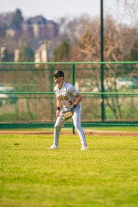 A young baseball player in uniform prepares to field on a sunny day at the ballpark. Green grass and a chain-link fence backdrop.