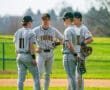Four baseball players in gray jerseys discussing strategy on the field under a clear blue sky.