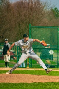 A baseball pitcher in gray Tigers uniform winds up to throw a pitch on a sunny day at the baseball field.
