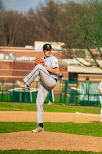High school baseball pitcher in grey uniform prepares to throw on the mound during a sunny game day.