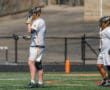Two lacrosse players in white uniforms on the field, one adjusting gear and the other in a ready stance.