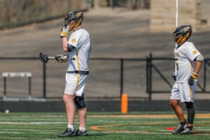 Two lacrosse players in white uniforms on the field, one adjusting gear and the other in a ready stance.
