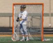 Lacrosse goalie in uniform stands in front of an orange net, ready for action on the field during a sunny day.