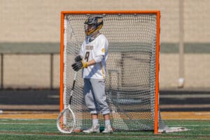 Lacrosse goalie in uniform stands in front of an orange net, ready for action on the field during a sunny day.