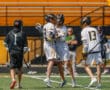 Players celebrating a goal during a high school lacrosse match on a bright, sunny field.