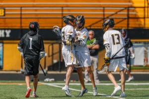Players celebrating a goal during a high school lacrosse match on a bright, sunny field.