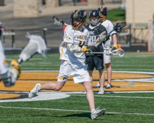 Lacrosse player in white uniform sprinting on the field, preparing to pass the ball during a game.