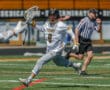 Lacrosse player from the Tigers team runs dynamically, preparing to take a shot during a game, with a referee present.