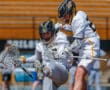 Two lacrosse players in action, one jumping over the other, showcasing teamwork and athleticism during a match.