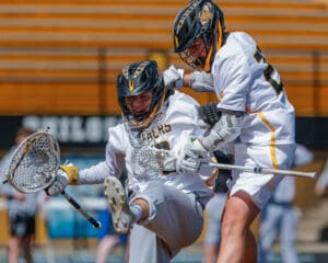 Two lacrosse players in action, one jumping over the other, showcasing teamwork and athleticism during a match.
