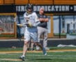 Lacrosse player in white uniform running on field, showcasing action during a game, with spectators in the background.