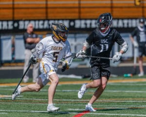 Two young lacrosse players compete fiercely on a bright, sunny field during a high-energy game.