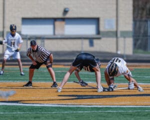 Two lacrosse players prepare for a face-off on the field, while an official watches closely in the background.