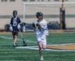 Player in white lacrosse uniform dodges while holding a stick on a sunny field during a match; teammate in background.