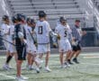 Lacrosse players in white uniforms celebrate during a game, with a referee observing the action on the field.