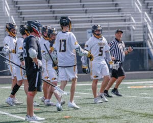 Lacrosse players in white uniforms celebrate during a game, with a referee observing the action on the field.