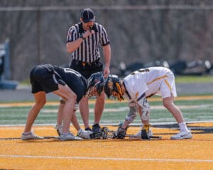 Two lacrosse players face off on a field with a referee overseeing the game, ready for the match to begin.