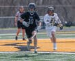 Two lacrosse players sprinting on a field during an intense game, with a referee in the background.