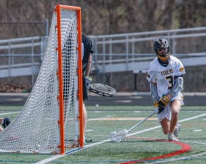 High school lacrosse player in action, wearing a Tigers jersey, attempting to score against the goalie.