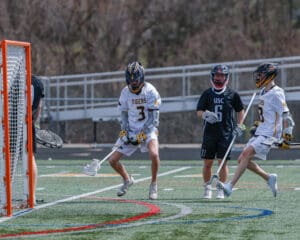 Lacrosse players in action during a game, focused on scoring while defending the goal on a sunny day.