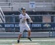 Player in white lacrosse uniform catches a ball during a game on a field with North Allegheny Tigers signage.