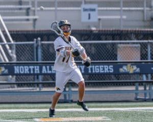 Player in white lacrosse uniform catches a ball during a game on a field with North Allegheny Tigers signage.
