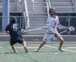 Two lacrosse players compete on a field, one preparing to dodge and pass while the other defends closely.