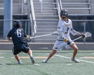 Two lacrosse players compete on a field, one preparing to dodge and pass while the other defends closely.