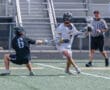 Lacrosse players in action: one defending, one attacking with a stick, while a referee observes during a game.