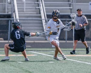 Lacrosse players in action: one defending, one attacking with a stick, while a referee observes during a game.
