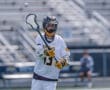 Lacrosse player in action, wearing a helmet and jersey, throwing a ball on a field with bleachers in the background.