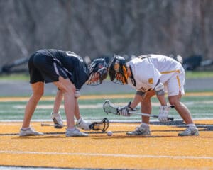 Two lacrosse players prepare for a faceoff on a bright, orange turf field during a game.