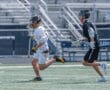Two lacrosse players sprinting on a field, one in white uniform and the other in black, during a game.