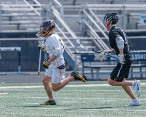 Two lacrosse players sprinting on a field, one in white uniform and the other in black, during a game.