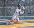 Young lacrosse player in white uniform dodges while carrying a lacrosse stick on a sunny field.