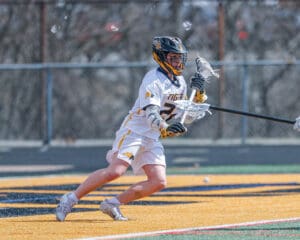 Young lacrosse player in white uniform dodges while carrying a lacrosse stick on a sunny field.