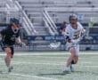 Two lacrosse players competing on the field, with one in a black uniform and the other in white.