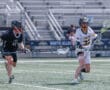 Two lacrosse players compete on a field, one in a black uniform and the other in a white Tigers jersey.