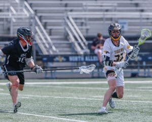 Two lacrosse players compete on a field, one in a black uniform and the other in a white Tigers jersey.