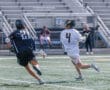 Two lacrosse players compete on a field, one in a black jersey chasing the other in white, showcasing athleticism and teamwork.