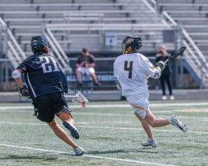 Two lacrosse players compete on a field, one in a black jersey chasing the other in white, showcasing athleticism and teamwork.