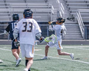 High school lacrosse players in action, one throwing the ball while two opponents defend on the field.