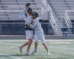 Two young lacrosse players celebrating a goal, embracing on the field with excitement and joy.
