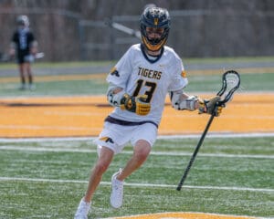 Lacrosse player in a Tigers jersey #13 dribbles the ball on the field during a game, showcasing athleticism and focus.