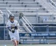 Lacrosse player in white uniform throws a ball during a game on a sunny day, showcasing skill and teamwork.