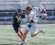 Two lacrosse players compete on the field; one in a black jersey is defending while the other, in white, advances with the ball.