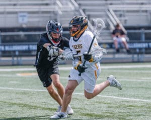 Two lacrosse players compete on the field; one in a black jersey is defending while the other, in white, advances with the ball.