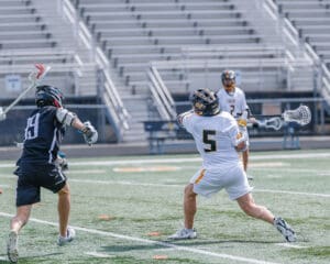 Lacrosse players in action on the field; one player passes while another defends. Competitive sports moment captured.