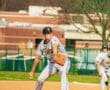 High school baseball pitcher in gray uniform winds up to throw a pitch on a sunny day at a baseball field.